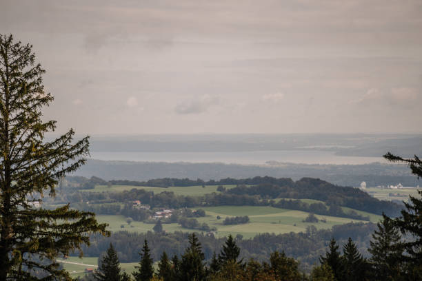 Bavaria, Germany - September 15, 2024 - A scenic view of a Bavarian landscape with rolling hills, forests, fields, and a distant lake under a cloudy sky.