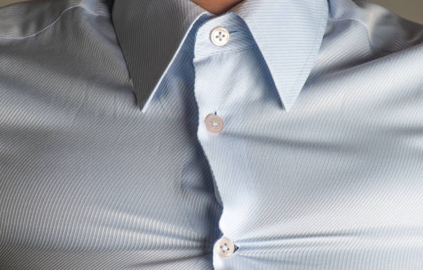 Close-up of a light blue business shirt on an overweight male, showing visible strain on the fabric. The buttons are tightly pulled, and the shirt appears wrinkled due to the tight fit, emphasizing the tension and discomfort. This image captures themes related to body image, tight clothing, weight issues, and wardrobe malfunctions.