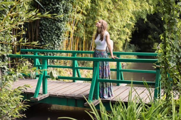 Young woman is exploring a beautiful garden. She is standing on a small bridge over a stream, admiring the the beauty of the nature.\nShot with Canon R5