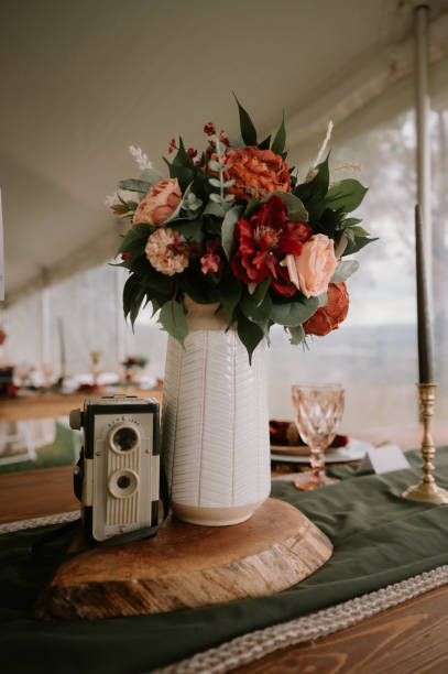 Large white vase overflowing with autumn florals sitting on a fall themed table scape with an olive green table runner, candle, and vintage camera. A close up of fall details on an autumn themed table scape. A tall white vase is filled with fall colored flowers-oranges, reds, pinks- with greenery. Next to the vase there is a silver vintage camera. The table scape consists of an olive green table runner, green candle stick and elegant glassware. pink memorial candle with flowers stock pictures, royalty-free photos & images