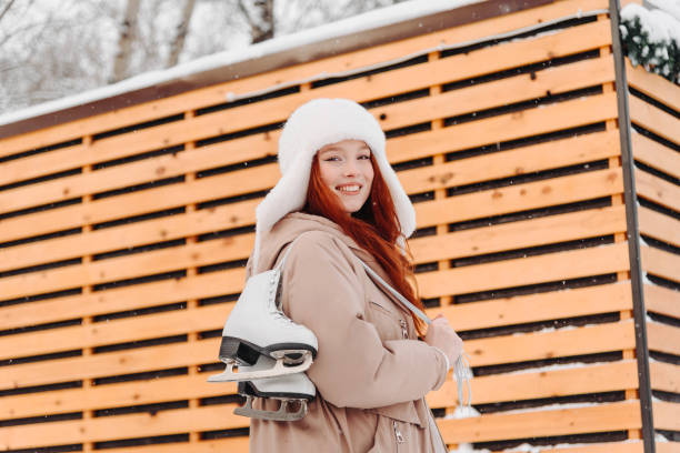 young pretty caucasian redhead smiling girl in white and beige winter clothes skating in ice rink in winter cloudy snowy day, with skates on shoulder, looking straight to camera