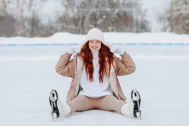 young pretty caucasian redhead girl in white and beige winter clothes skating in ice rink in winter cloudy snowy day, sitting funny, smiling widely