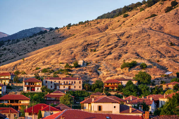 The village of Agios Germanos in Greece, nestled at the foot of arid hills, illuminated by the warm light of sunset, with traditional stone houses and red-tiled roofs.