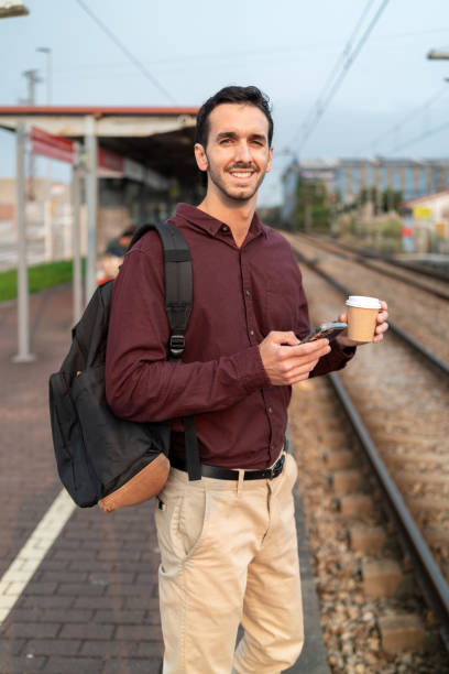 istock Smiling young professional standing on train platform, holding coffee and using smartphone 2175357101