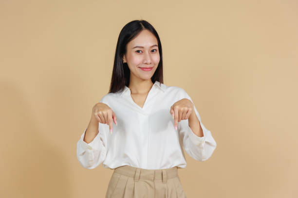istock Smiling Woman Pointing Downwards with Both Hands, Beige Background 2175383563
