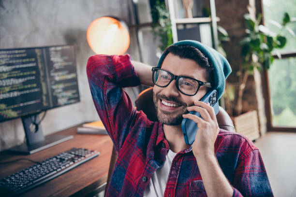 Photo of positive cheerful coder dressed hat glasses smiling talking apple samsung modern gadget indoors workplace workstation loft