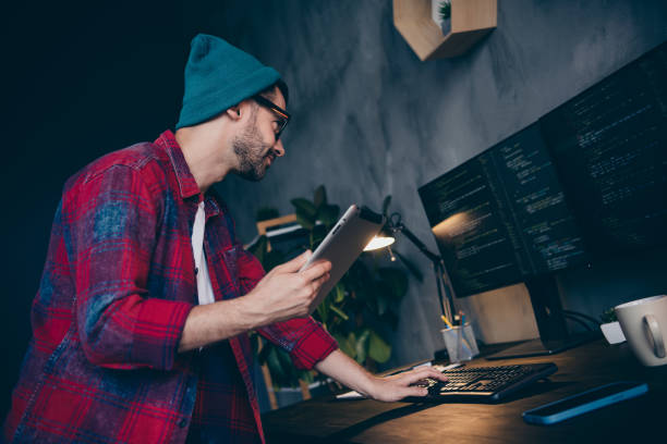 istock Photo of serious busy coder dressed hat glasses connecting ereader to modern gadget indoors workplace workstation loft 2175407984