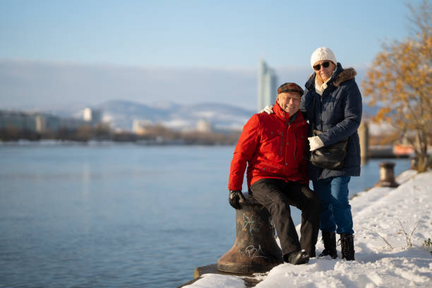 happy senior couple enjyoing winter day outdoors in snow at riverbank of european city on cold sunny day, having short rest after walk, smiling at camera