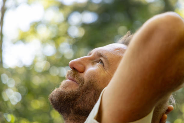 A close-up side profile of a man relaxing outdoors, looking peaceful with a forest background. Sunlight filters through the trees, creating a serene, natural ambiance.