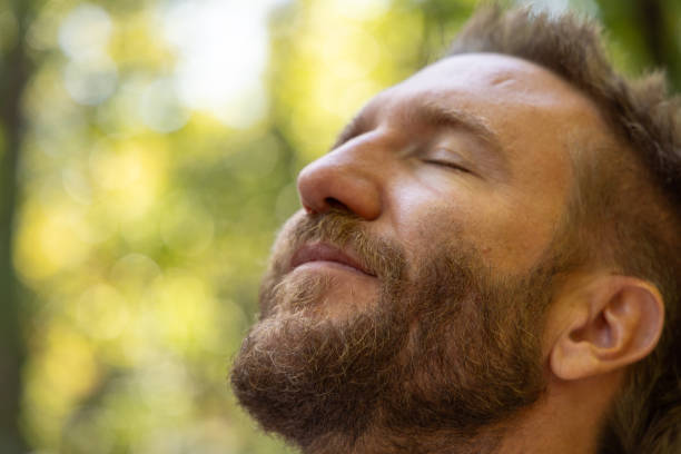 Close-up of a man meditating with eyes closed, enjoying peaceful nature. Sunlight filters through trees, creating a serene atmosphere of relaxation.