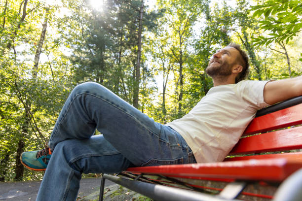 Smiling man sitting on a red bench in a sunlit forest, legs stretched out, enjoying a peaceful moment in nature. Sunlight filters through the trees, creating a serene atmosphere.