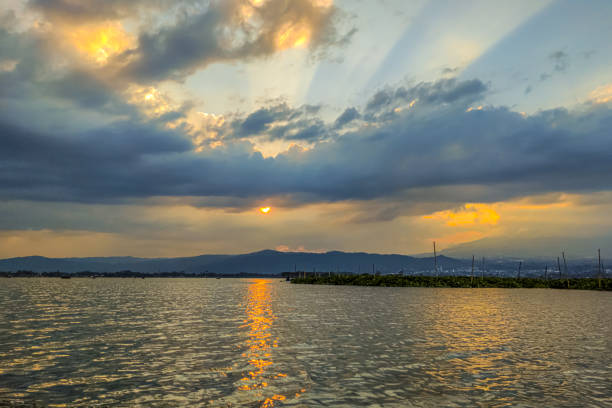 Serene view of Rawa Pening lake at sunset. Sky ablaze vibrant colors, dramatic clouds and golden sunlight streaming through. Calm lake reflects the sky and mountains in distance, creating mirror-like effect. Atmosphere is peaceful and stunning, showcasing beauty of nature at its finest.
