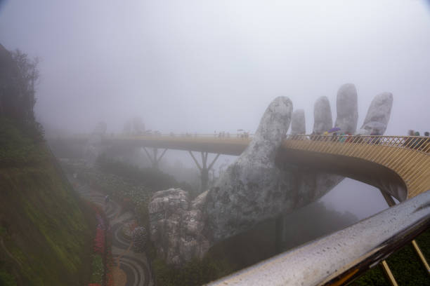 Da Nang City, Vietnam - December 13, 2023: Tourists walk on the mist-covered Golden Bridge in Ba Na Hills, supported by giant hands. The bridge is located 1,400 meters above sea level with hill scenery. The Golden Bridge is a 150-metre-long (490 ft) pedestrian bridge designed to connect the cable car station with the gardens and provide a scenic lookout for tourists. The bridge loops around, and has two giant hands, constructed of fiberglass and wire mesh, designed to appear like stone hands that support the structure.