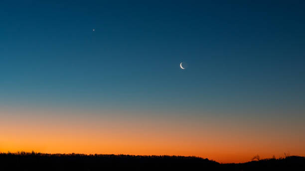 A crescent moon and the Northern Star are visible against a deep blue sky at twilight, with an orange glow on the horizon above the silhouetted mountains. Serene early dawn landscape. stock photo