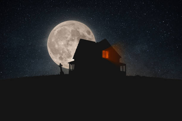 An old dilapidated house with light from the window stands on a hill with a cemetery at night with stars in the full moon. happy halloween wallpaper. Dark halloween, creative idea