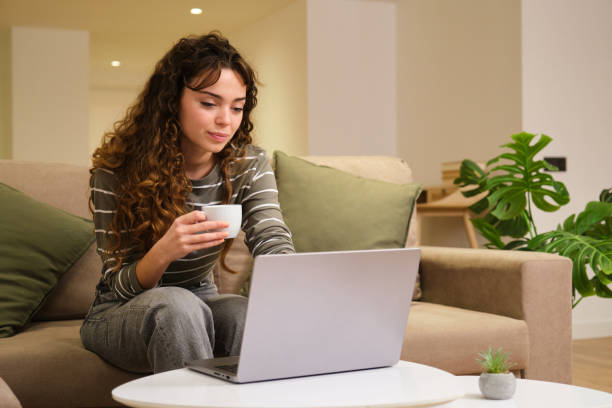 istock Young woman sitting on sofa using laptop and drinking coffee at home 2175492689