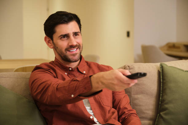 istock Handsome man sitting on sofa is watching television and holding a remote control 2175493276