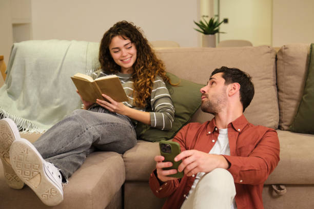 istock Young woman reading book while boyfriend using smartphone at home 2175493499