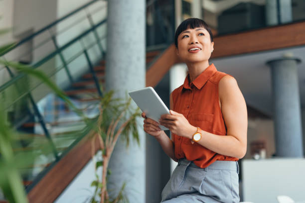 Smiling Japanese Professional Woman Using Tablet in Modern Office Setting Confident Asian woman in a orange blouse holding a tablet and smiling in a contemporary office environment, representing success and technology in the workplace. new idea office stock pictures, royalty-free photos & images