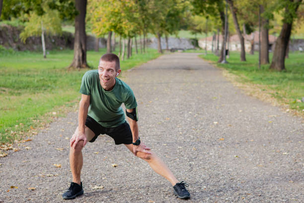 Happy athletic mid adult man doing gentle everyday exercise outdoors in public park, as part of his mental wellness and self-care activities. He is wearing sports clothes and smartwatch, and he is stretching after jogging.