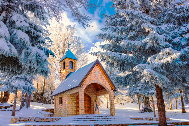 A wooden chapel sits nestled in a snow-covered forest. Frost-laden trees frame the scene, creating a warm yet serene atmosphere despite the cold surroundings. This charming winter scene invites a feeling of peaceful reflection.