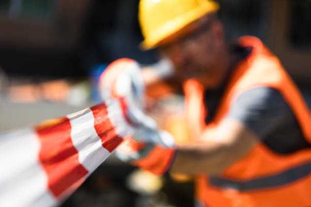 A construction worker in safety gear handles caution tape while overseeing a construction site. The bright sun illuminates the bustling urban area around him.