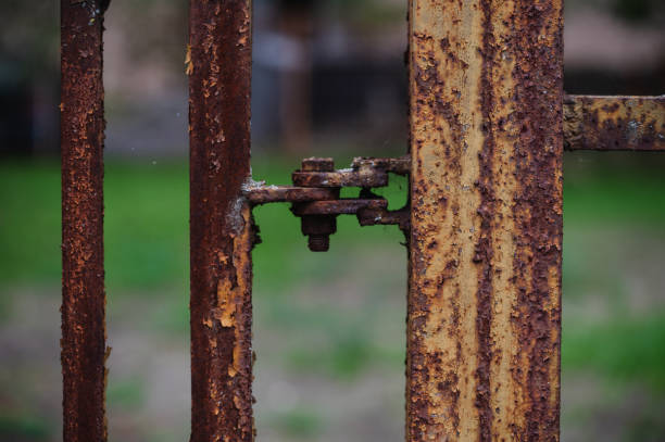 A detailed close-up of a rusty metal fence hinge, showcasing the corroded texture and weathered appearance. The aging metal contrasts with the blurred green background, emphasizing the passage of time.