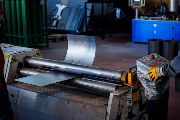 depicts a worker handling a flat sheet of metal as it is fed into a rolling machine. The worker is wearing red gloves and appears to be carefully aligning the sheet for processing. The rolling machine, which has two large metal rollers, is designed to bend or shape the metal sheet into a curved form. The setting is an industrial workshop, and the background shows various equipment and machinery, including a control panel and storage racks. The environment sa metal fabrication or manufacturing facility specializing in custom metal forming processes.