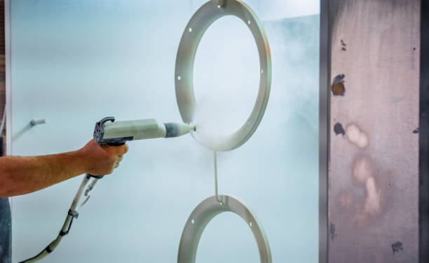a close-up of a powder coating process being applied to a metal ring or circular component. The worker is holding a powder coating gun, which emits a fine spray of powder paint onto the surface of the ring. The powder coating creates a smooth and even layer on the metal object. The environment appears to be a spray booth designed to ensure a clean and controlled coating application. The background is plain and industrial, emphasizing the focus on the coating process.