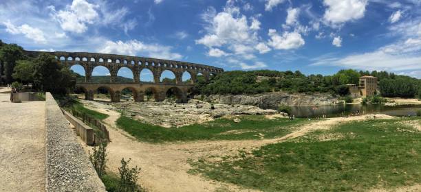 Panoramic wide shot of tourists admiring a surviving and iconic Roman Aqueduct traversing a scenic river in Pont du Gard, France