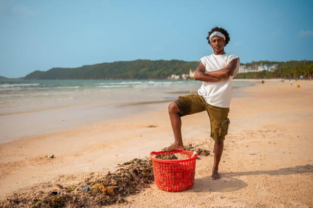A self-assured young adult male stands by the ocean, arms crossed, dressed casually in front of a serene tropical beach, with a red basket at his feet.