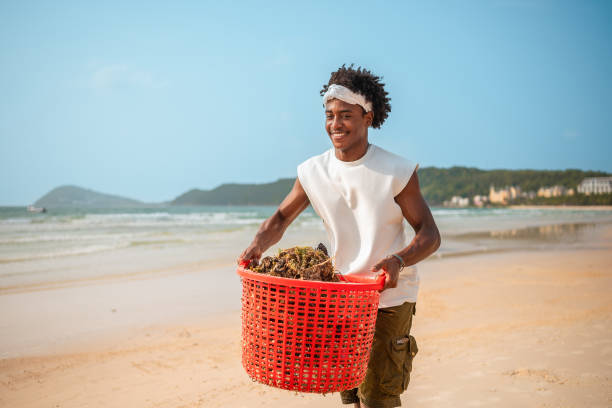 A cheerful young Black man actively engages in environmental conservation by collecting trash with a red basket on a sunlit tropical beach, dressed in casual summer attire.