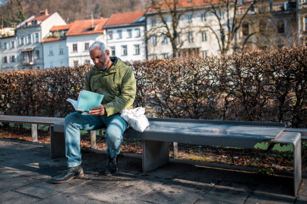 An elderly Hispanic male sits absorbed in reading a magazine on a park bench. The outdoor setting is tranquil, with lush greenery in the background, suggesting a peaceful urban park environment. He is dressed in casual wear appropriate for a mild climate.