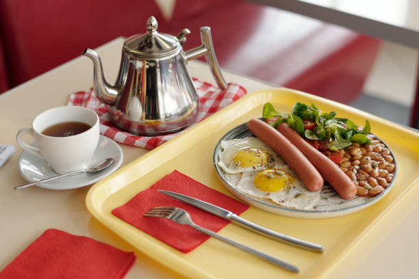 A satisfying and hearty breakfast plate, typically served in a diner or cafe setting. The dish features a large, fluffy pancake dusted with powdered sugar and topped with a swirl of whipped cream. On the side are two pieces of crisp, savory bacon and a browned breakfast sausage link. The plate is completed with two sunny-side-up eggs and a generous serving of crispy, seasoned home fries (diced potatoes). A side plate holding two slices of buttered toast is visible in the background.