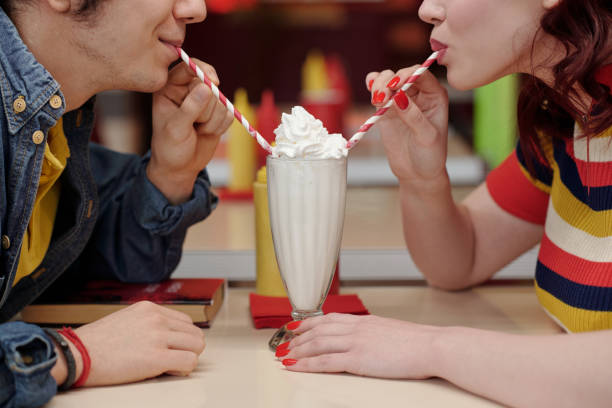 Two people enjoying a milkshake together, each sipping through striped straws while sitting closely in a cozy diner setting, creating an intimate and friendly atmosphere