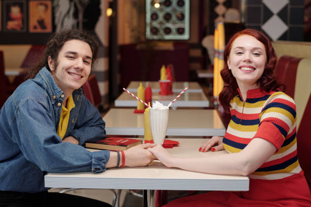 Young couple happily sitting at a retro diner table while holding hands and enjoying milkshakes place has a nostalgic ambiance
