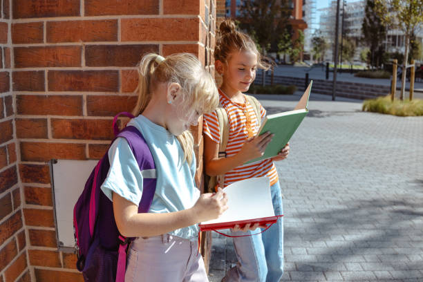 Two young girls stand outside reading notes in their notebooks, enjoying an educational moment in a city environment on a sunny day.