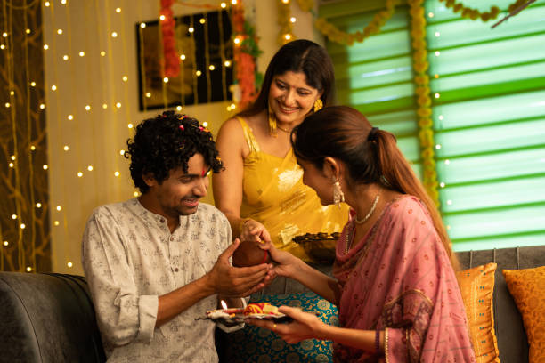 Woman looking at sister giving religious offerings to brother during Bhai Dooj Smiling young woman looking at sister giving coconut to smiling brother while celebrating Bhai Dooj festival in living room at home bhai tika stock pictures, royalty-free photos & images