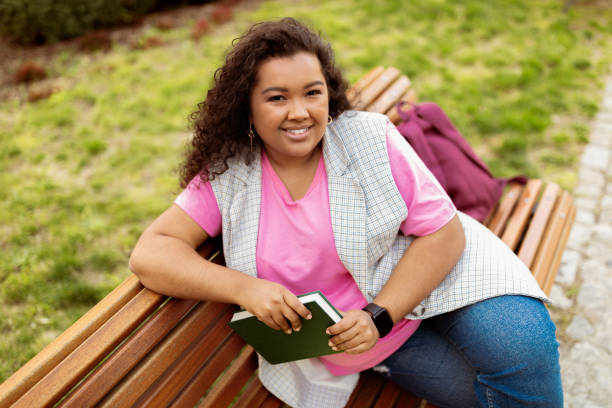 istock Young woman enjoying a sunny day outdoors while reading a book on a bench 2175603336