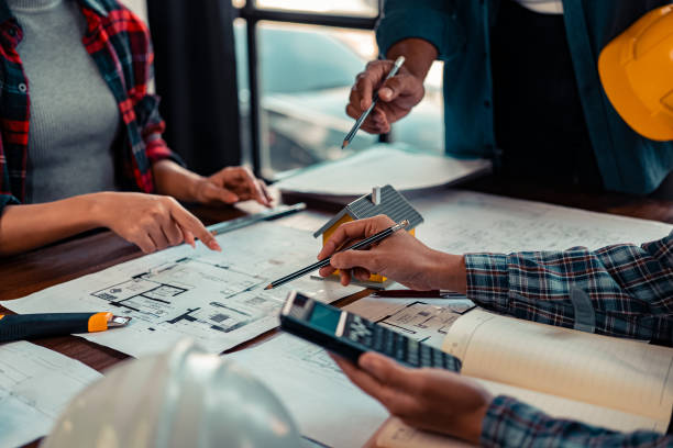 Architects and engineers point to blueprints of architectural drawings inside a building to discuss construction plans, with hard hats and architectural documents scattered around. Close-up.