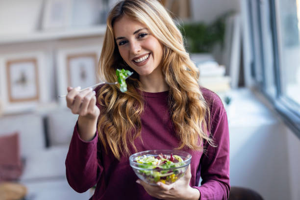 istock Pretty smiling woman eating healthy salad while looking at camera standing in the kitchenPortrait of pretty smiling woman eating healthy salad while looking at camera standing in the kitchen 2175632905
