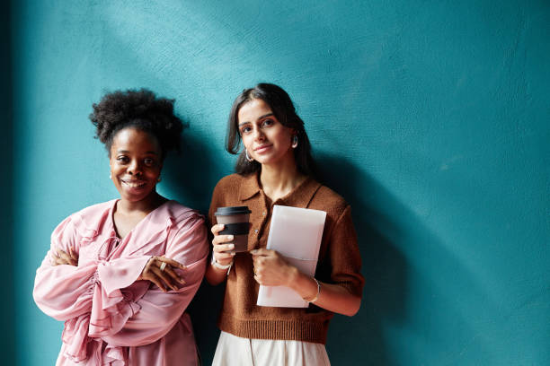 Two Businesswomen in Office Clothing Smiling at Camera while Standing against Blue Wall