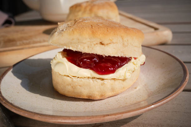 Stock photo showing close-up view of an English afternoon tea scene, featuring a blue and white striped Cornish ware plate containing golden, plain scones filled with clotted cream and strawberry jam. The scones are filled in both the Cornish way with jam spread on the scone first, followed by a dollop of clotted cream, and the Devonshire way with cream first with a spoonful of jam on top. Social debate concept.
