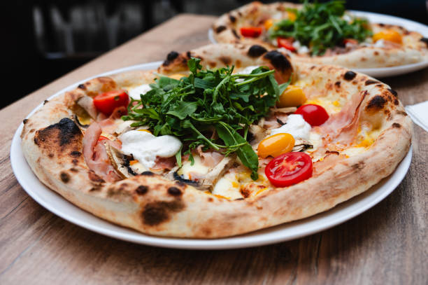 A delicious gourmet pizza topped with fresh arugula, colorful cherry tomatoes, slices of ham, and dollops of cheese. The pizza has a perfectly baked, golden-brown crust, served on a white plate on a wooden table.
