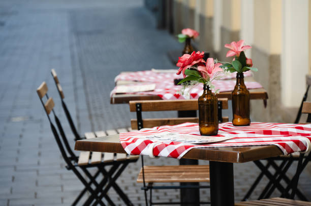 A charming outdoor cafe setup with wooden tables and chairs, each table adorned with a red checkered tablecloth and a small vase of pink flowers. The cozy street-side ambiance invites passersby to sit and enjoy a peaceful meal or coffee.
