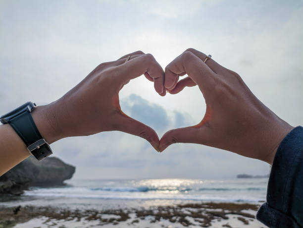 The happy couple shows a heart symbol by forming a heart with their hands, meaning love, against the backdrop of a tropical beach and ocean. Sign of love on an exotic tropical beach.