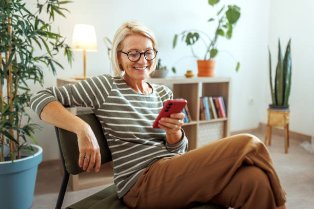 istock Smiling mature woman using smart phone while sitting in chair at home 2175665940