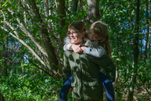 One girl giving another a piggyback in sun-dappled woods