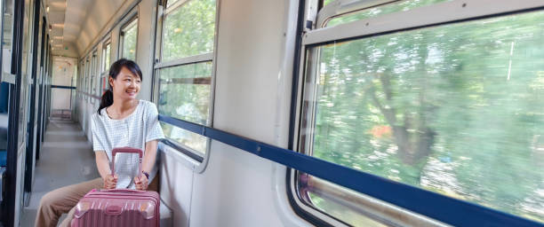 A middle-aged Asian woman travels alone on the train. She drags a suitcase on the aisle of the train and looks at the scenery outside the window.
