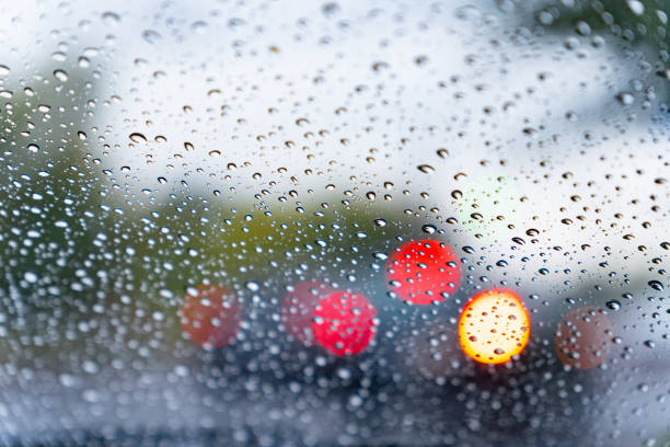 Raindrops cling to a car windshield, distorting the view of bright red and yellow lights from vehicles moving in the rainy city. The atmosphere feels moody and reflective.